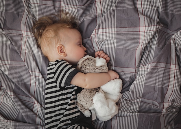 Portrait of a cute six-month-old blond boy sleeping on a gray bed with a toy in his hands