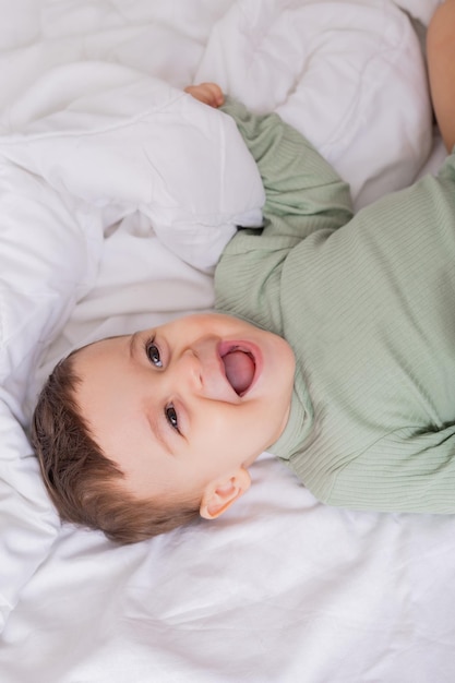 Portrait of a cute smiling baby in a green cotton bodysuit lying on a white bed on his back high