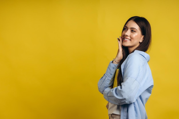 Portrait cute smiling classic dressed student girl looking directly to the camera and pointing with her index finger to the left side isolated over yellow background copy space