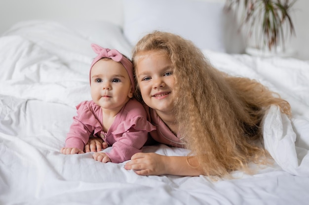 Photo portrait of a cute smiling curly blonde girl and a baby lying in bed