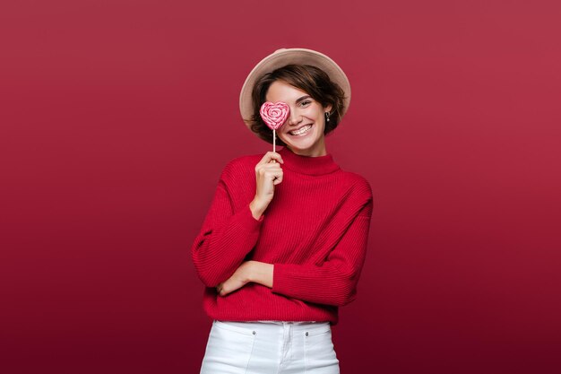 Portrait of cute smiling french woman wearing hat holding lollipop candy covering one eye