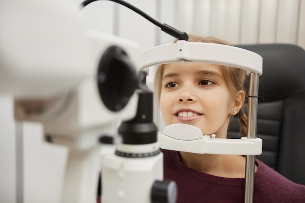 Portrait of cute smiling girl looking into refractometer during vision test