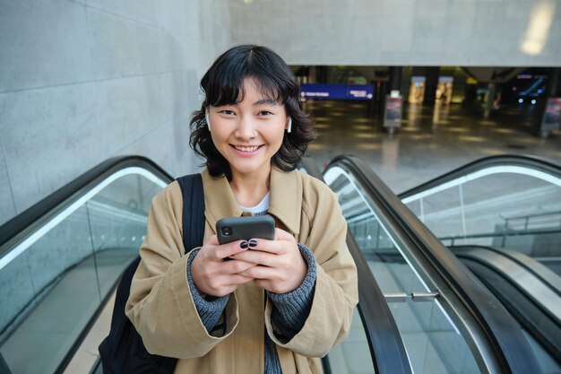 Portrait of cute smiling korean girl goes up escalator listens music in wireless earphones and uses