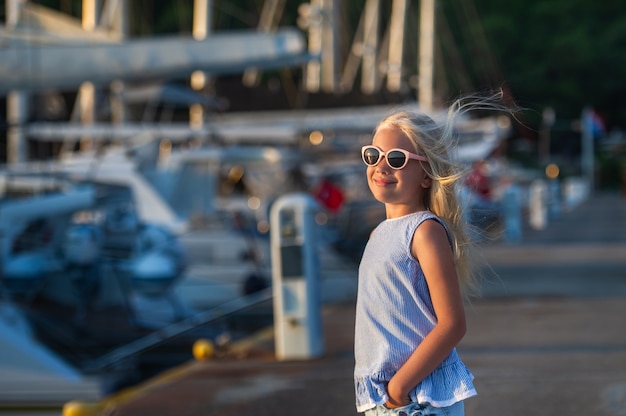 안경을 쓴 귀여운 웃는 어린 소녀의 초상화. 반바지를 입은 소녀와 바다에서 해질녘 파란색 티셔츠. 터키