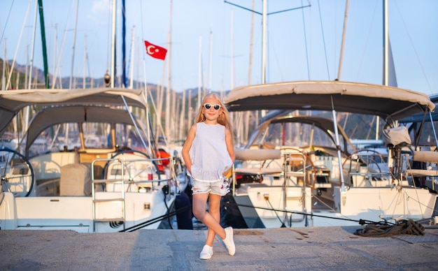 Portrait of a cute smiling little girl with glasses on a pier with yachts.A girl in shorts and a blue T-shirt at sunset near the Yachts.Turkey.