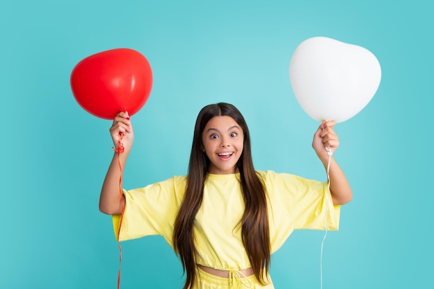 Portrait of a cute smiling teenager girl with a red heart\
balloon over blue background isolated excited face cheerful\
emotions of teenager girl