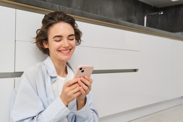 Portrait of cute smiling woman sitting on floor at home with crossed legs looks at mobile phone chat