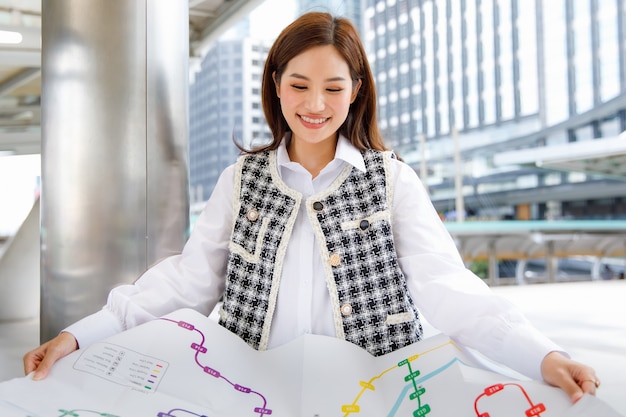 Portrait of a cute smiling young adult asian woman in smart
casual black and white fashion clothes leaning against the pole,
holding a paper metro map and looking at camera with a blurred
background