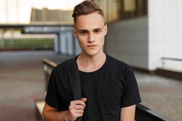 Photo portrait of a cute stylish young man in a black t-shirt with a sports black backpack with a stylish hairstyle on the background of the building