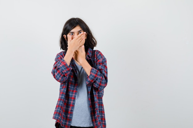 Portrait of cute teen girl holding hand on mouth in checked shirt and looking scared front view