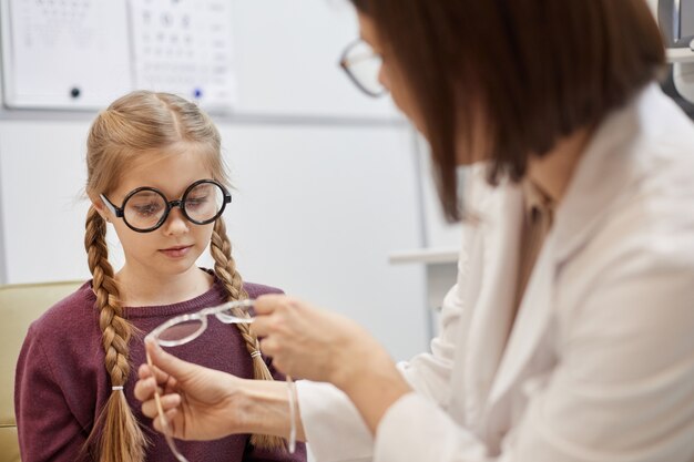 Portrait of cute teenage girl trying on glasses during vision test in modern ophthalmology clinic