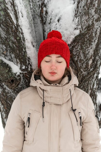 Portrait of cute thoughtfully girl with closed eyes Pretty young woman in red hat and mittens stands near snowcovered tree