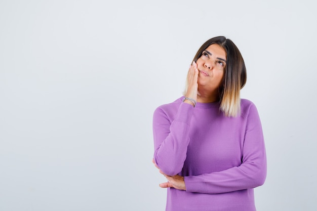 Portrait of cute woman leaning cheek on hand, looking up in purple sweater and looking thoughtful front view