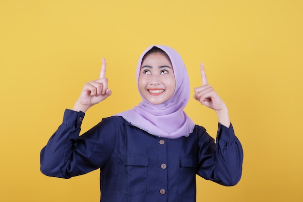 Portrait of cute woman pointing up in casual cloth wearing hijab on isolated yellow background