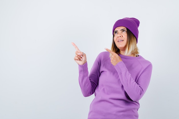 Portrait of cute woman pointing at upper left corner, looking up in sweater, beanie and looking dreamy front view