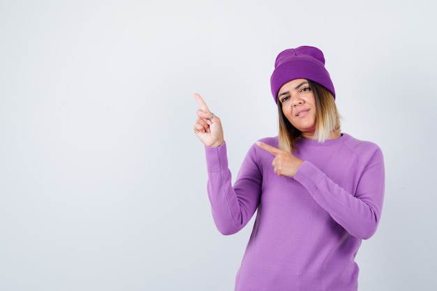 Portrait of cute woman pointing at upper left corner in sweater, beanie and looking cheery front view