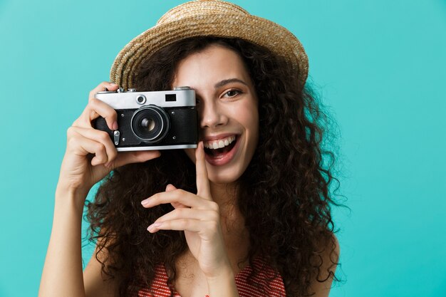 Portrait of cute woman wearing straw hat photographing on retro camera, isolated over blue wall