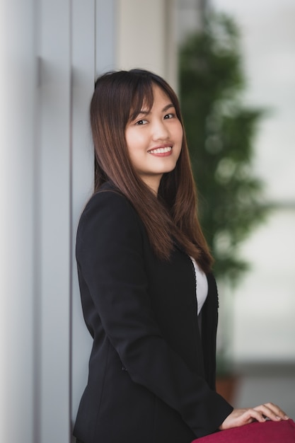 Portrait of cute and young Asian female entrepreneur or secretary in elegant outfit standing with friendly smile face beside windows in office hall and looking at camera