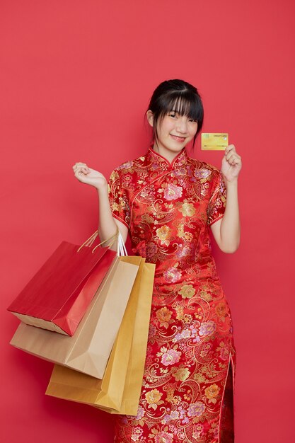 Portrait Cute young Asian woman wear Chinese dress with a shopping bag and credit card for Chinese new year on a red wall