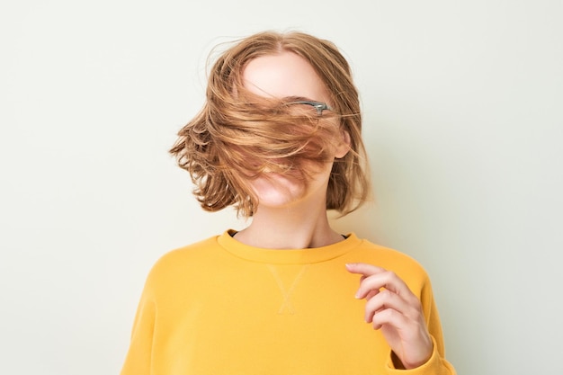 Portrait of cute young blond girl in glasses and yellow sweater shakes her head and plays with hair isolated on white studio background