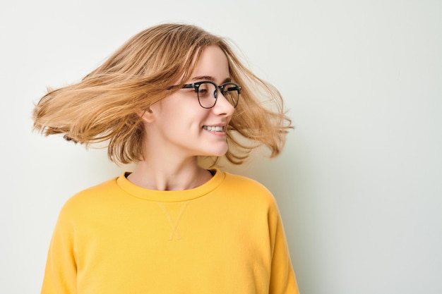 Photo portrait of cute young blond girl in glasses and yellow sweater shakes her head and plays with hair isolated on white studio background