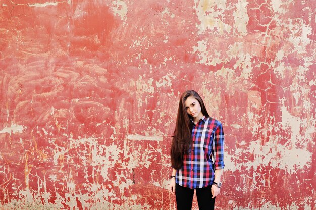 Portrait of cute young brunette girl in plaid shirt on red grunge wall surface. Copy space, space for text