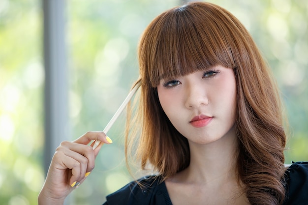 Portrait of  cute young and good looking  Asian brown hair woman smiling and playing pencil in her hand with relax and hpaay pose in front of green tree blurred background.