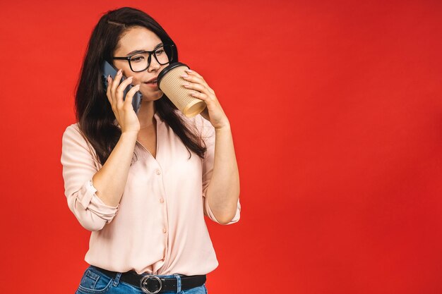 Portrait of cute young joyful business woman wearing casual using talking on mobile phone isolated over red background Drinking morning cup of coffee