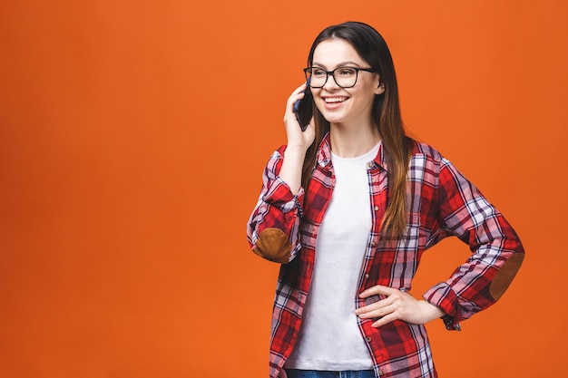 Portrait of cute young joyful woman with trendy hairstyle wearing casual, talking on mobile phone isolated over orange background.
