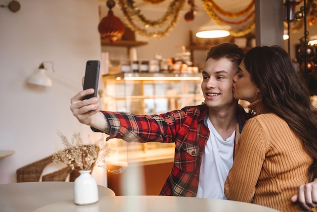 portrait of a cute young loving couple sitting in cafe indoors take a selfie by mobile phone.