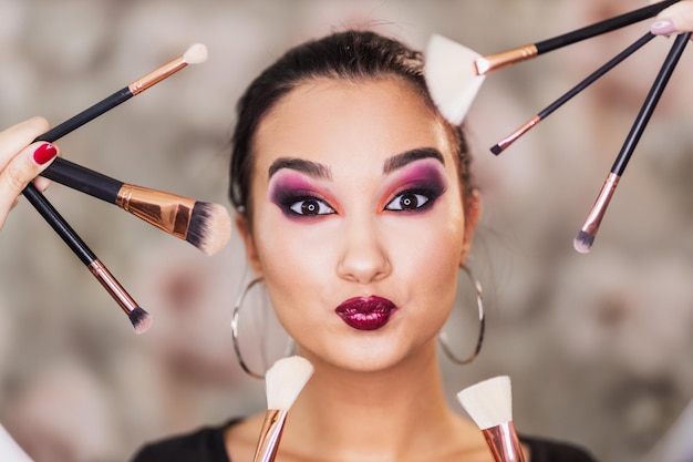 A portrait of a cute young woman surrounded by makeup brushes who is presenting a nice makeup on her face.