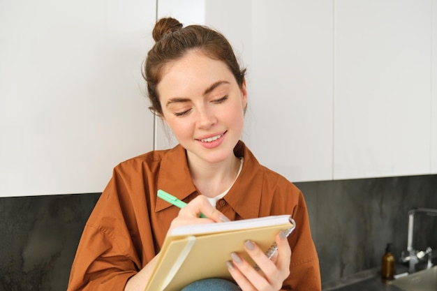 Portrait of cute young woman with notebook sitting in the kitchen and writing her to do list making