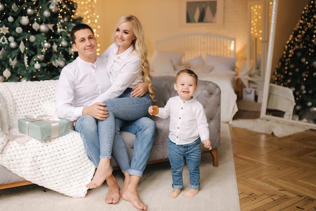 Portrait of dad, mom and son sitting on the floor at home near the Christmas tree, all are smiling. Denim and white shirts. Christmas family. Happiness.