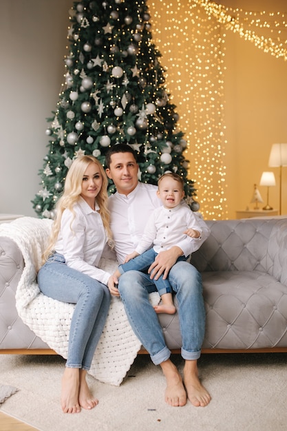 Portrait of dad, mom and son sitting on the floor at home near the Christmas tree, all are smiling. Denim and white shirts. Christmas family. Happiness.