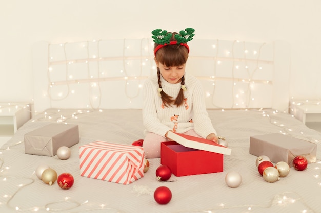 Ritratto di bambina dai capelli scuri che indossa un maglione bianco e corna di cervo cerchio di carnevale seduto sul letto e aprendo la scatola regalo, celebrando il natale e le vacanze invernali.