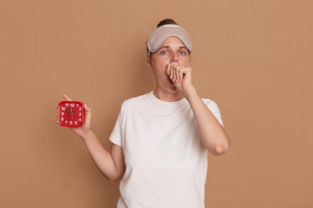 Portrait of dark haired young adult woman in cat sleeping eye mask yawning and covering mouth with hand holding red alarm clock isolated over brown background