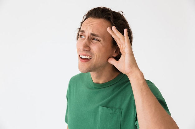 Portrait of deaf young man trying to hear something while keeping hand at his ear isolated over white wall