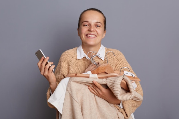 手にハンガーに服を持って立って、こぼれるような笑顔でカメラを見て灰色の背景の上に孤立して立っている携帯電話を保持している喜んでいる魅力的な女性の肖像画