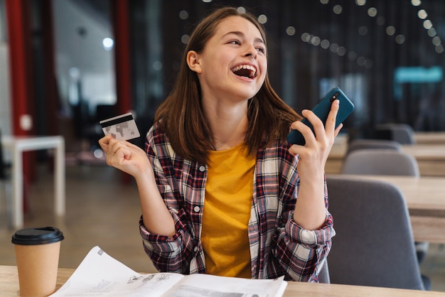 Photo portrait of delighted cute girl holding credit card and cellphone while doing homework in college library