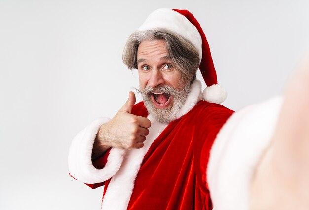 Portrait of delighted old man wearing Santa Claus red costume taking selfie with thumb up isolated on white