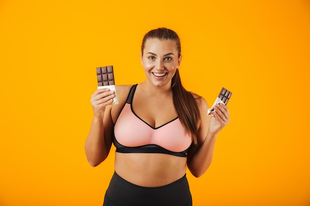 Portrait of a delighted overweight young woman wearing sport clothing standing isolated over yellow wall, eating chocolate