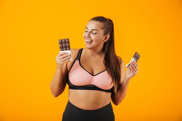 Portrait of a delighted overweight young woman wearing sport clothing standing isolated over yellow wall, eating chocolate