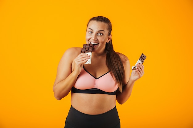 Portrait of a delighted overweight young woman wearing sport clothing standing isolated over yellow wall, eating chocolate