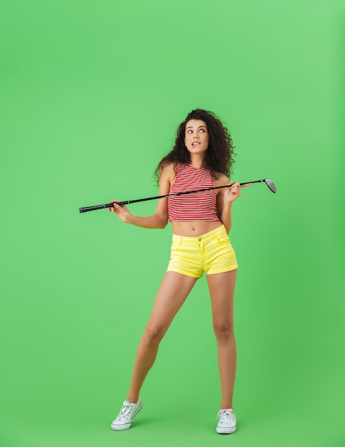 Портрет счастливой женщины 20 лет в летней одежде, держащей клюшку и играющей в гольф, стоя на зеленой стене