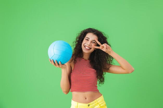 Portrait of delighted woman 20s wearing summer clothes smiling\
and holding volley ball while standing on green