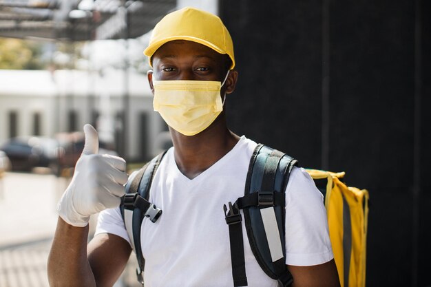 Portrait of delivery man with yellow cap in face mask and\
thermal backpack looking at camera