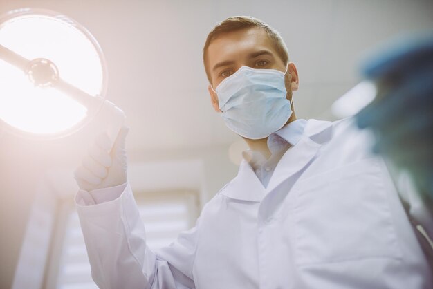 Portrait of dentist in medical mask adjusting lamp and holding dental tool in modern dental clinic