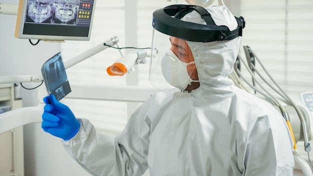 Portrait of dentist with face shield explaining holding dental x-ray to patient pointing on monitor during covid-19 pandemic. Orthodontic doctor wearing protection suit, coverall, mask and gloves