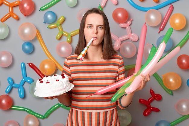 Portrait of depressed offended bored woman with brown hair wearing striped dress standing around multicolored inflated balloons celebrating birthday alone with cake and party horn being in bad mood