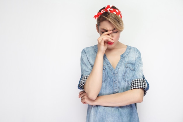 Portrait of depressed unhappy beautiful young woman in casual blue denim shirt with red headband standing holding head down, hopeless and crying. indoor studio shot, isolated on white background.